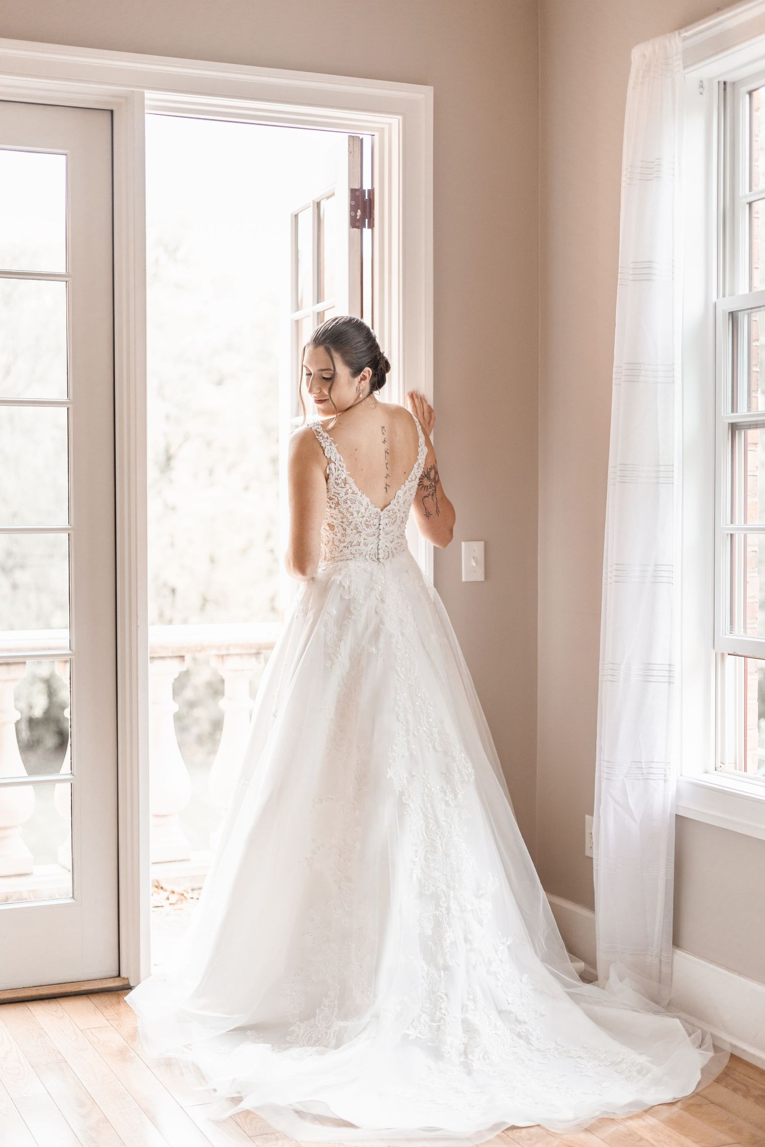 Bride in elegant lace ball gown with deep V-back standing by French doors in soft natural light.