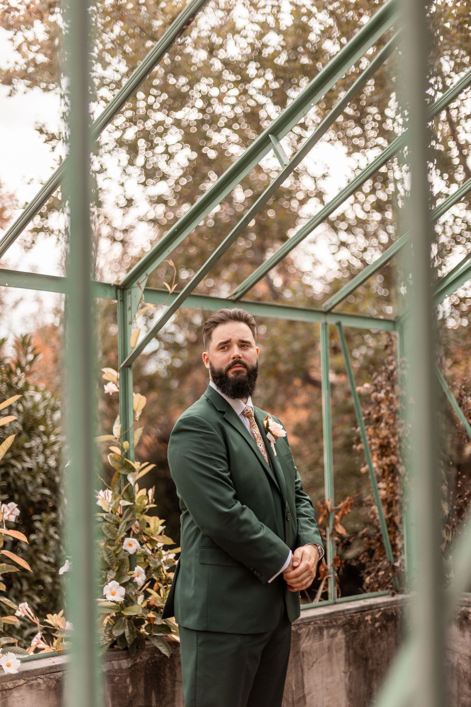 Dapper groom in dark green suit with boutonniere posing inside elegant greenhouse wedding portrait setting.