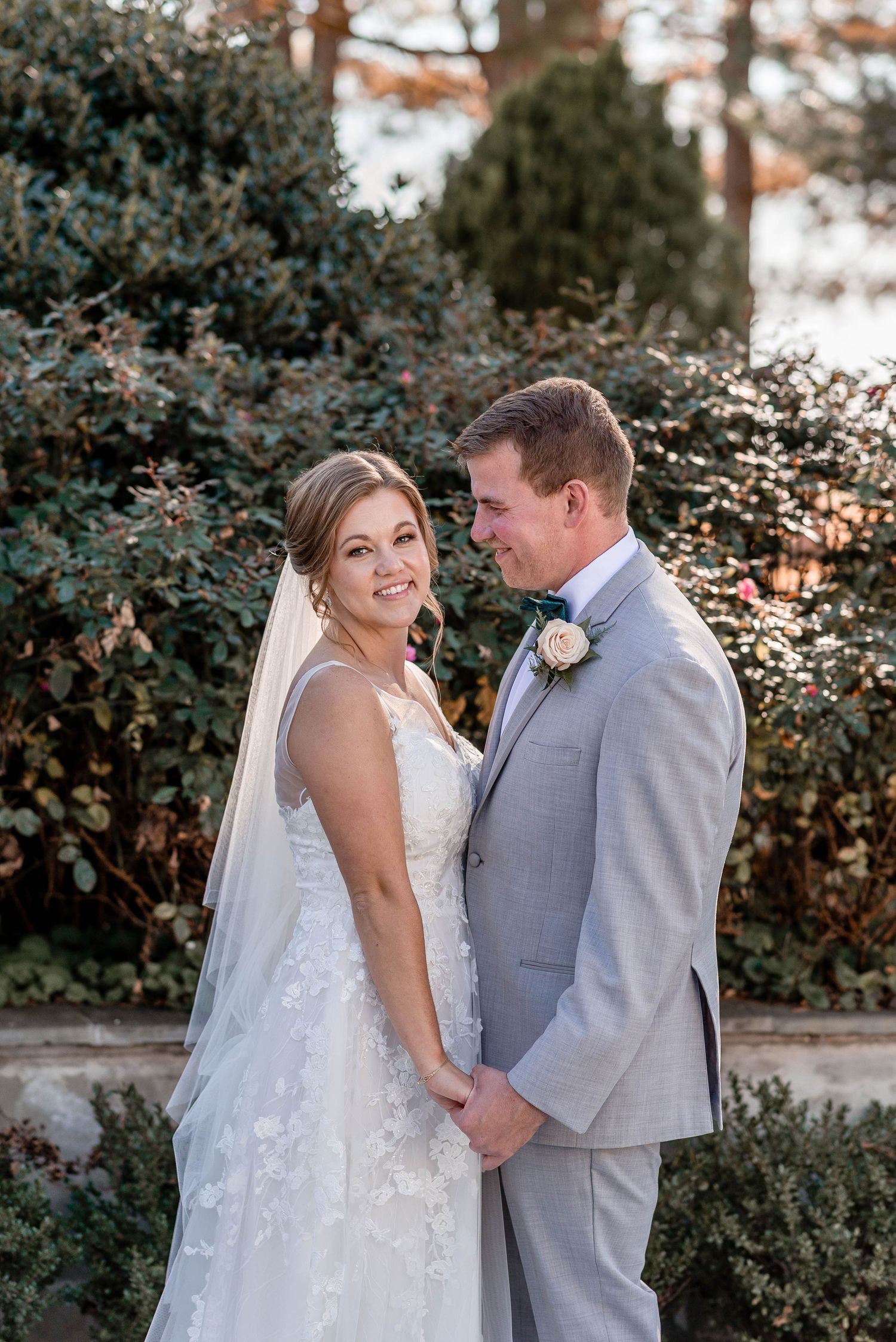 Bride and groom posing together in front of lush green bushes on wedding day.
