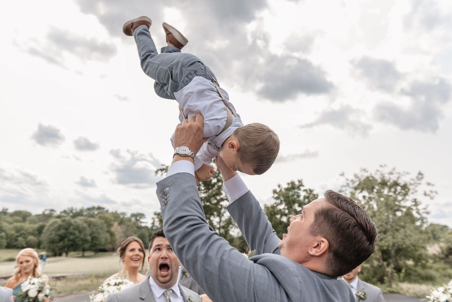 Excited groom in gray suit lifting laughing baby boy into the air at outdoor wedding.