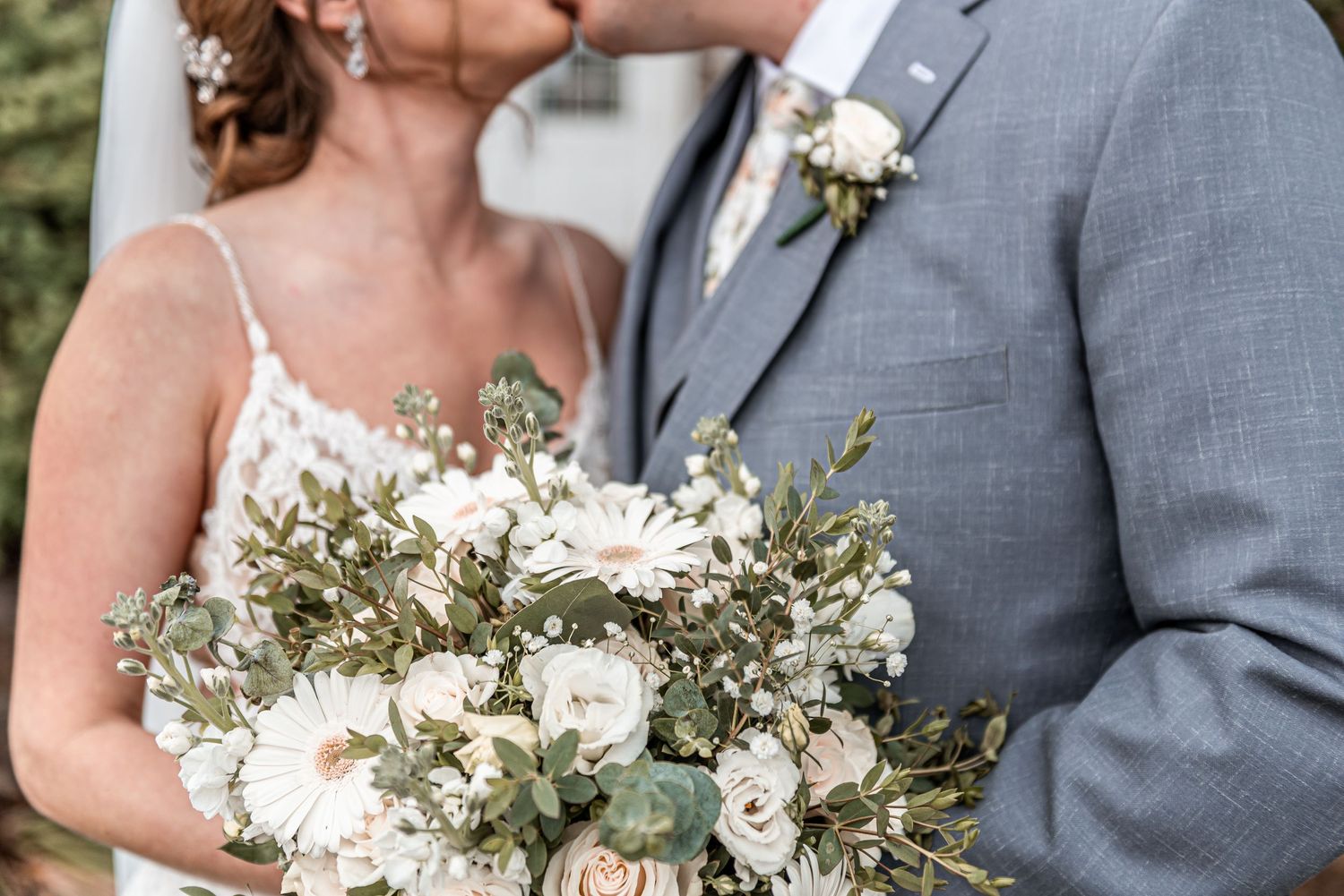 Bride and groom share a kiss while holding a lush white and greenery wedding bouquet.
