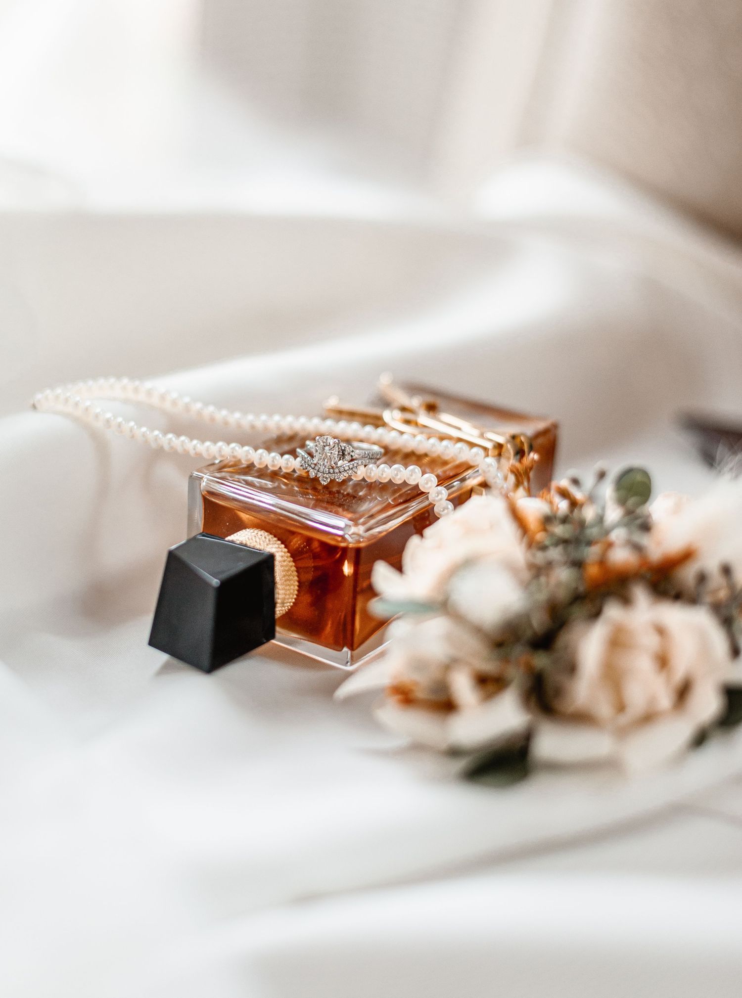 Wedding flat lay featuring a diamond ring, pearl necklace, amber perfume bottle, and soft floral arrangement on white silk.