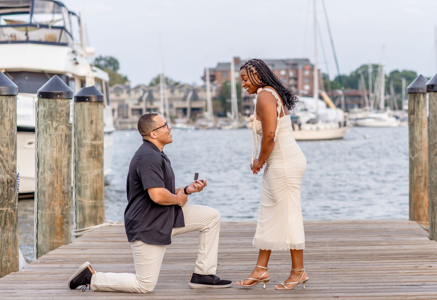 Man proposing on one knee to woman on a marina dock with boats in the background.