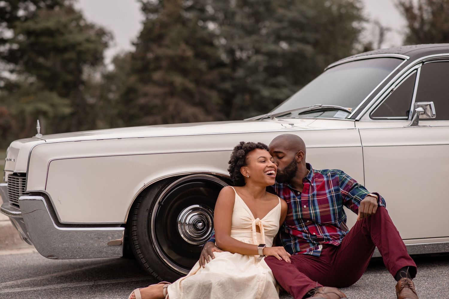 A happy couple sits on the pavement leaning against a classic white vintage car sharing a tender kiss.
