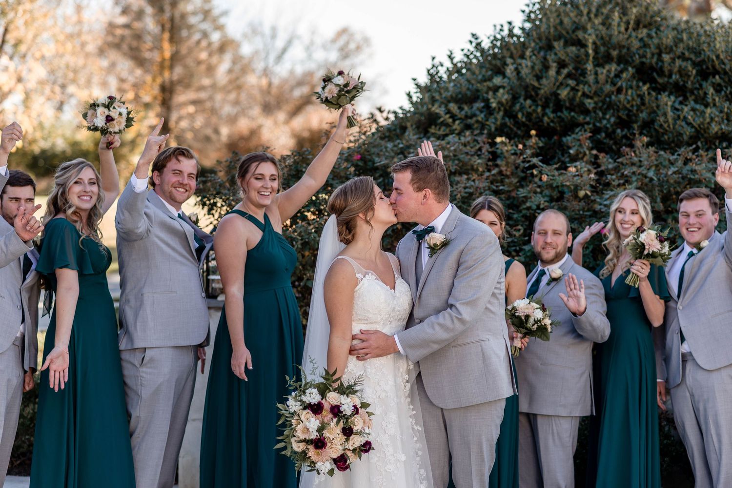 Wedding party celebrates as bride and groom kiss, bridesmaids in teal gowns and groomsmen in grey suits.
