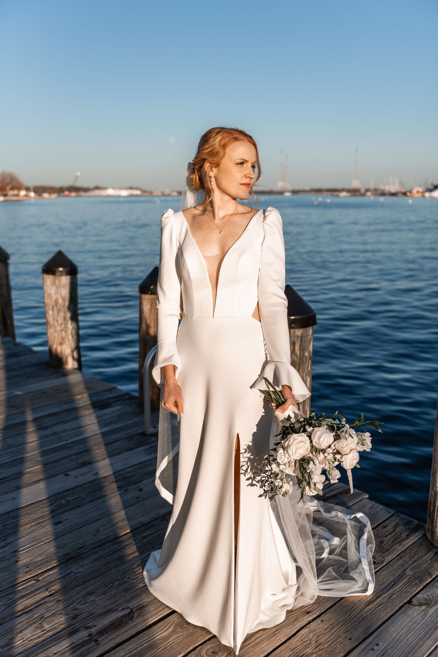 Elegant bride in a white long-sleeve gown with deep V-neck holds a bouquet on a sunny waterfront dock.