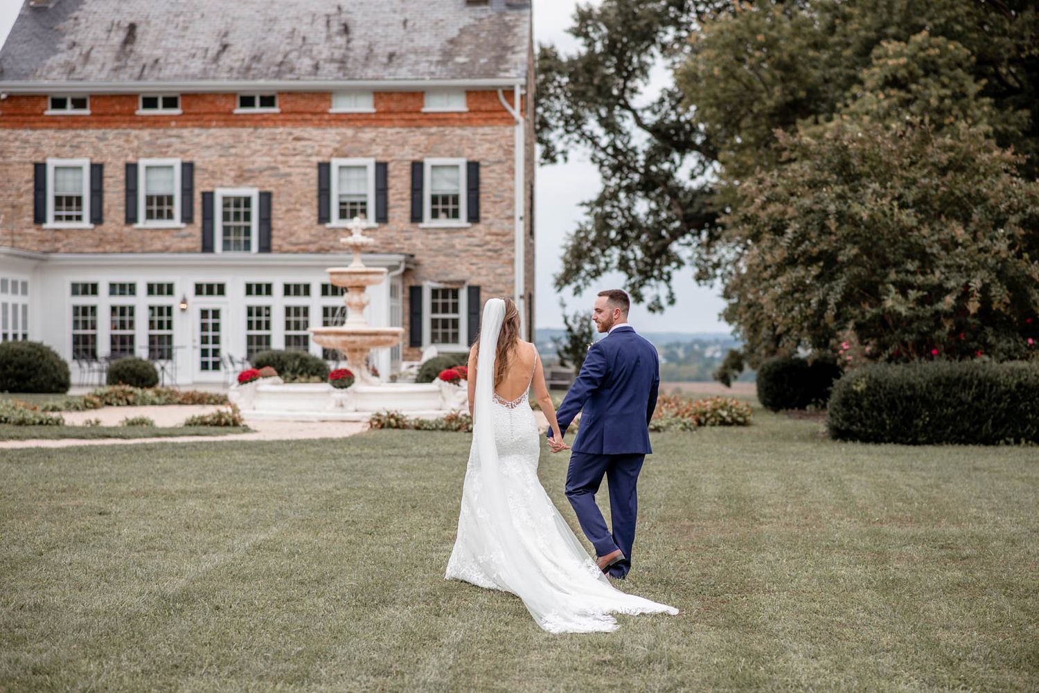 Bride and groom walk hand in hand across a lush green lawn in front of an elegant brick estate.