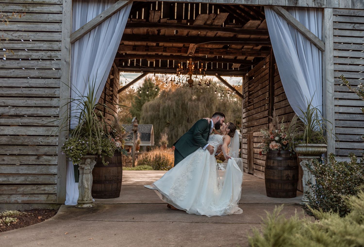 Bride and groom share a romantic kiss in a rustic barn doorway adorned with white drapes and barrel flower arrangements.