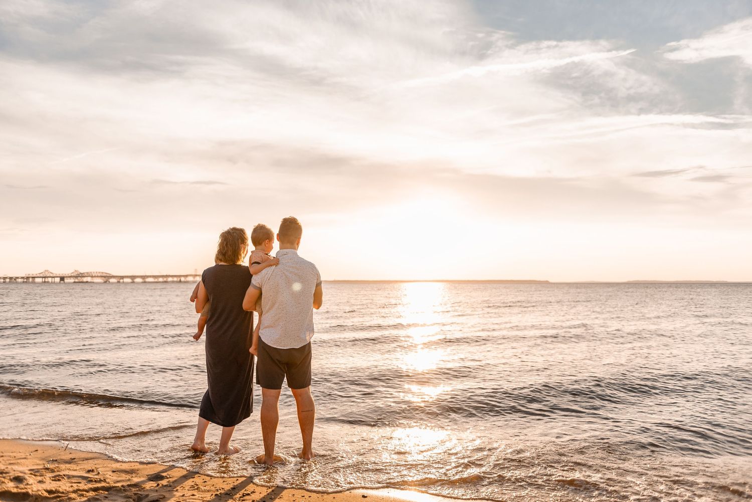 A family of three stands together on a sandy beach watching a breathtaking golden sunset over the ocean.