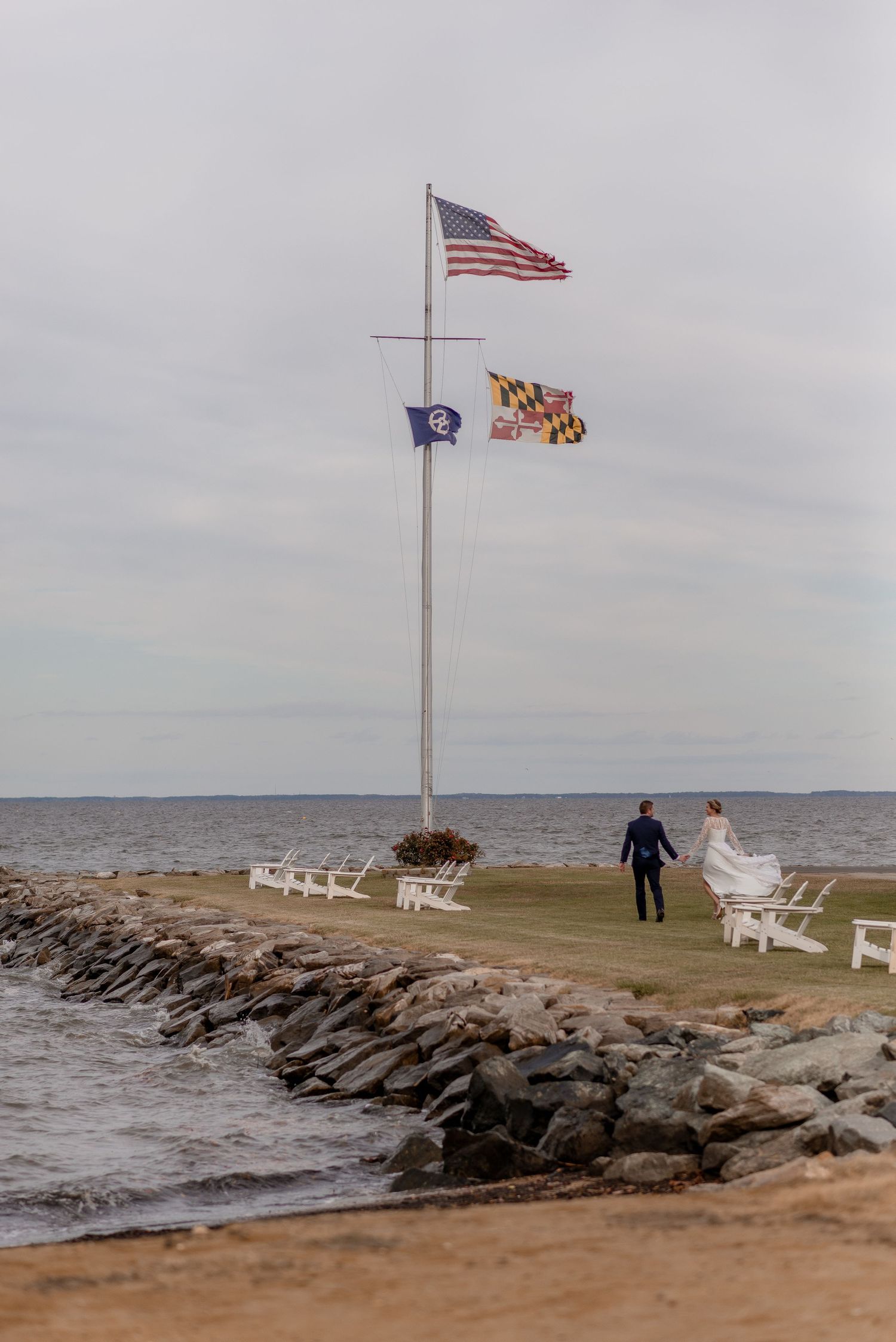 Newlyweds stroll along rocky shoreline near flagpole flying American and state flags.