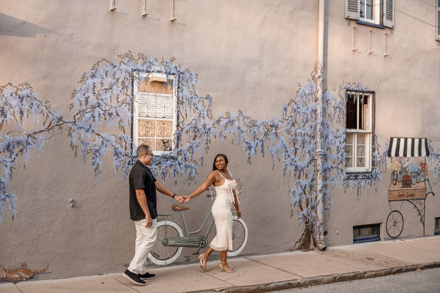 Couple holding hands walking past a blue wisteria mural on a charming street with a vintage bicycle.