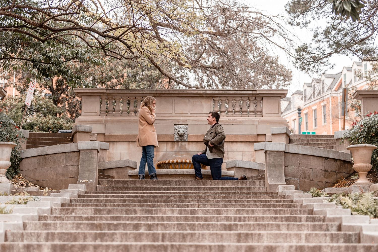 Man proposing on one knee on grand stone steps while woman reacts with surprise outdoors.