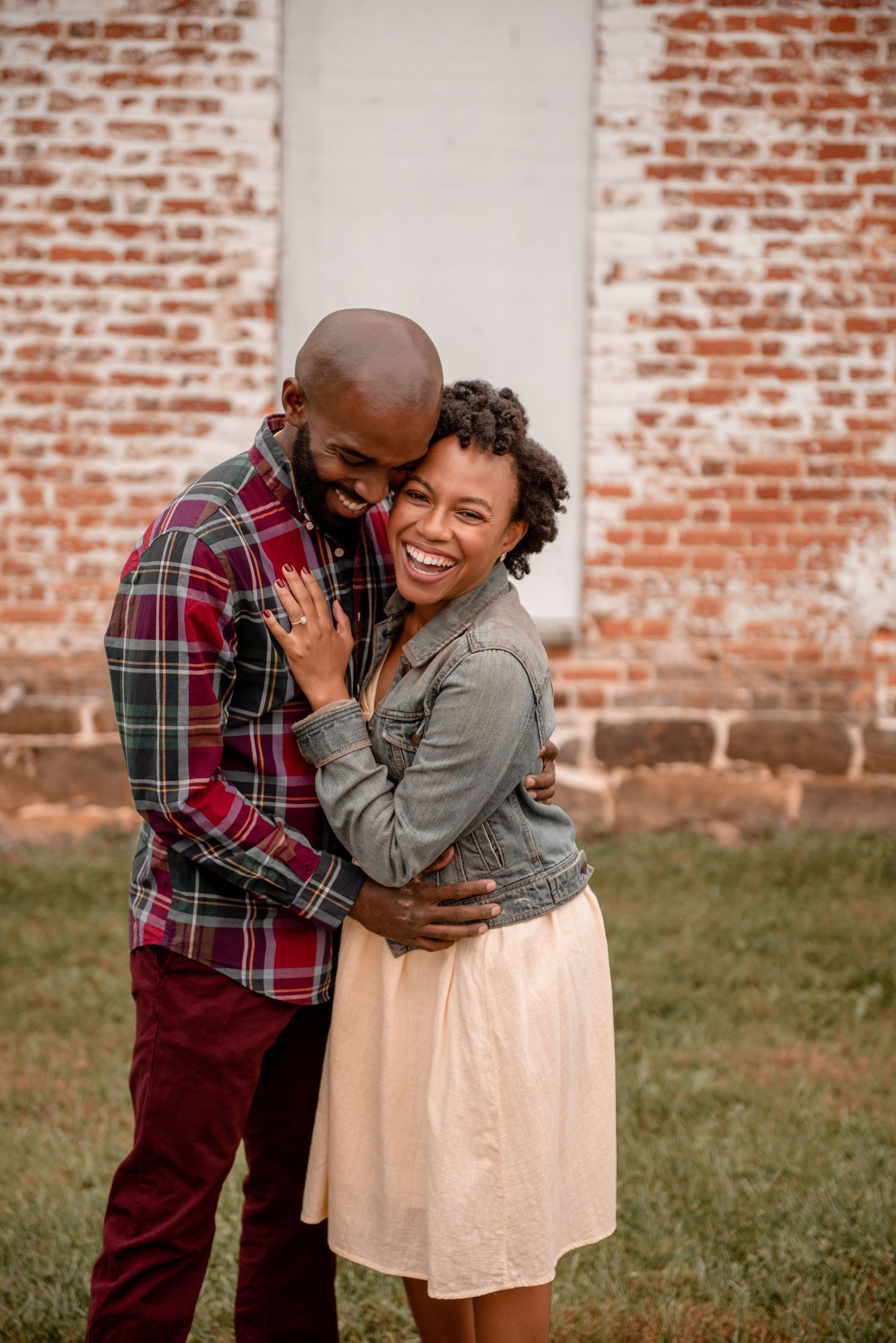 Joyful couple embracing and laughing in front of rustic brick wall during outdoor engagement session.