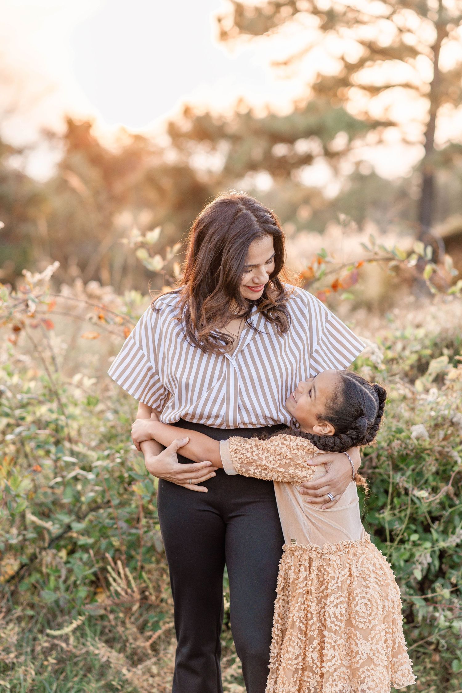 A woman in a striped blouse shares a warm hug with a young girl in a golden dress outdoors.