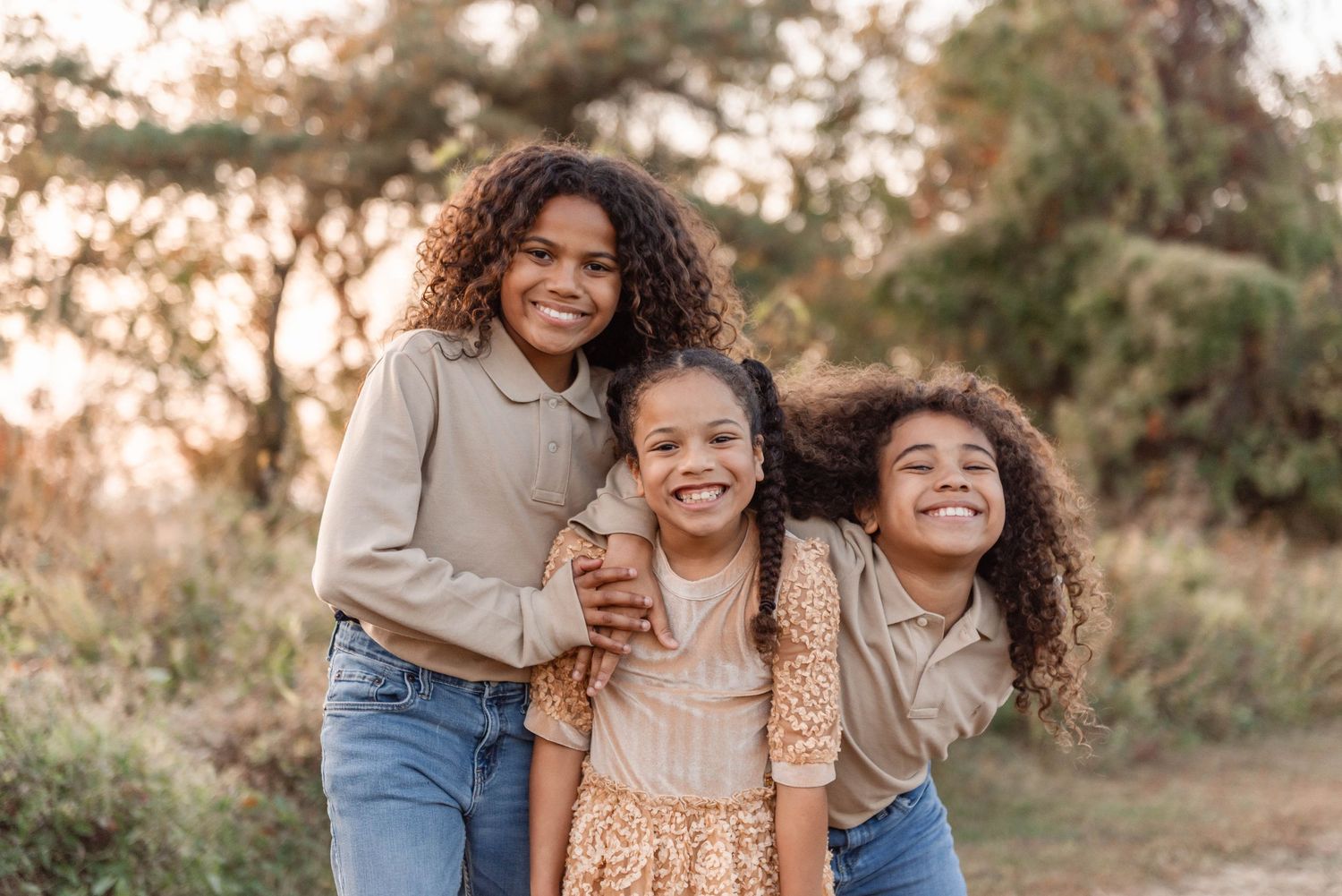 Three curly-haired sisters smiling and embracing outdoors during a golden autumn day.