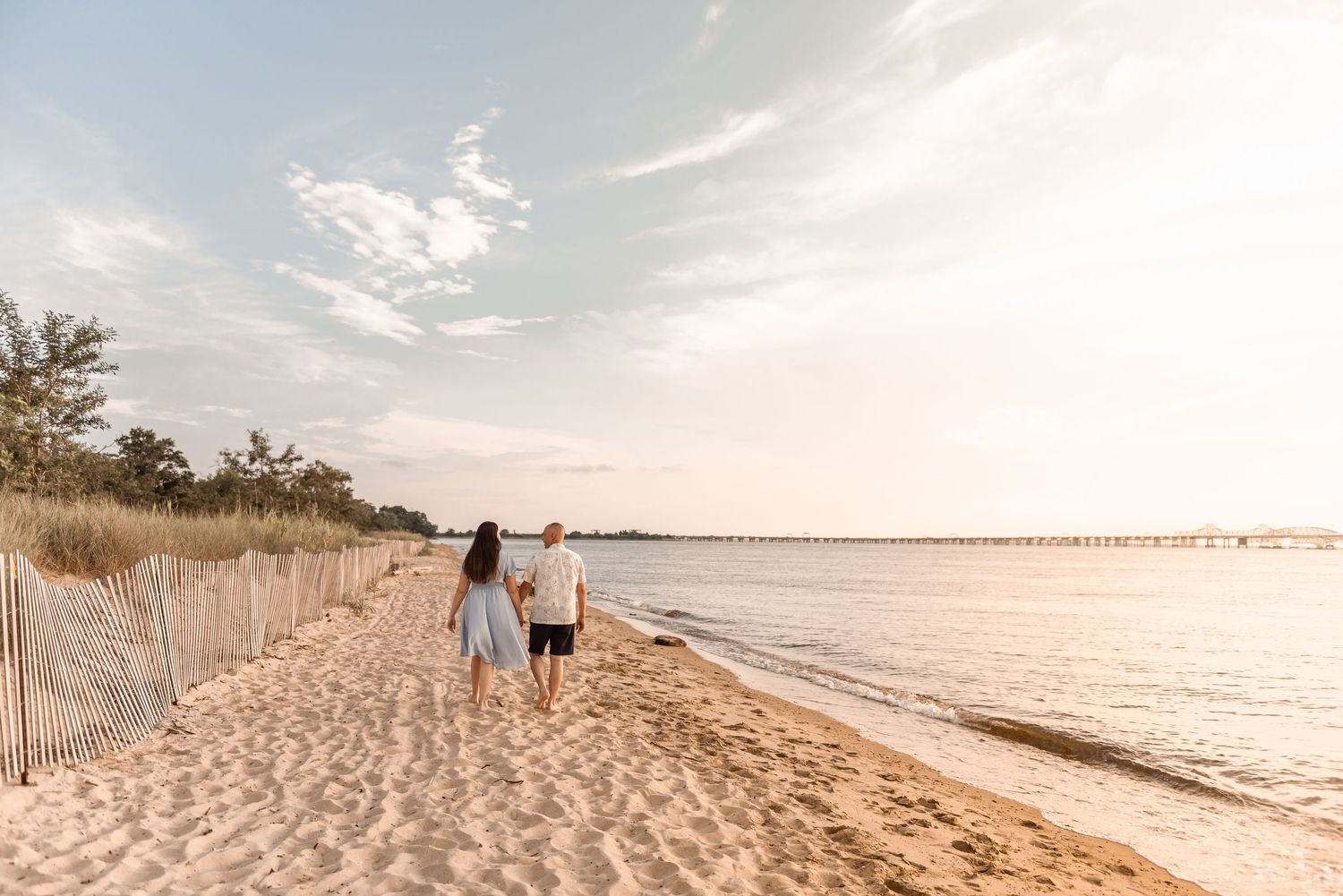Couple walking hand in hand along a sandy beach at golden hour with a pier in the distance.