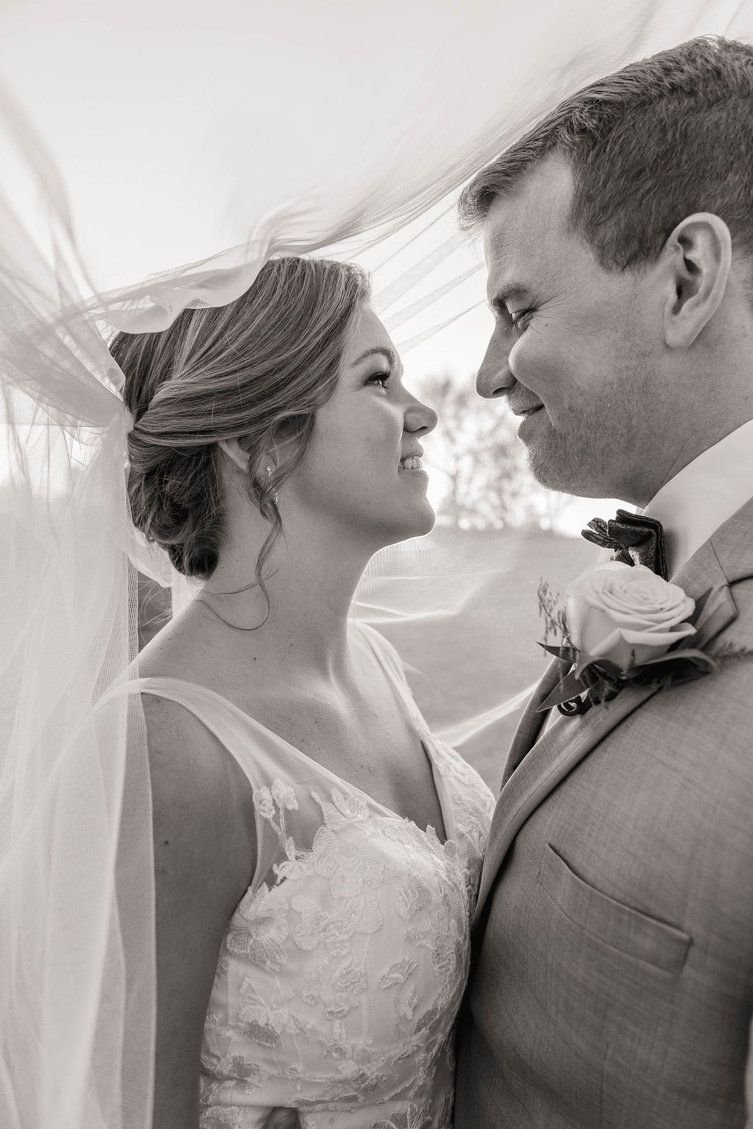 Black and white close-up of smiling bride and groom sharing an intimate moment beneath flowing veil.