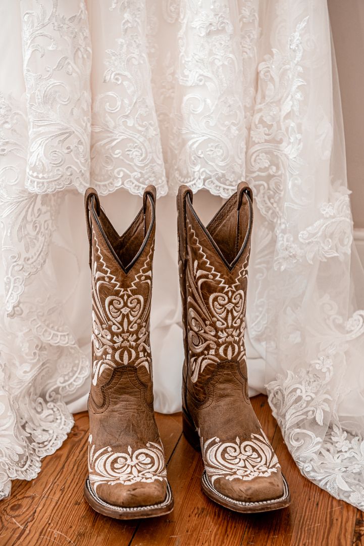 Brown and white embroidered cowboy boots displayed beneath a lace wedding dress hem on wood floor.