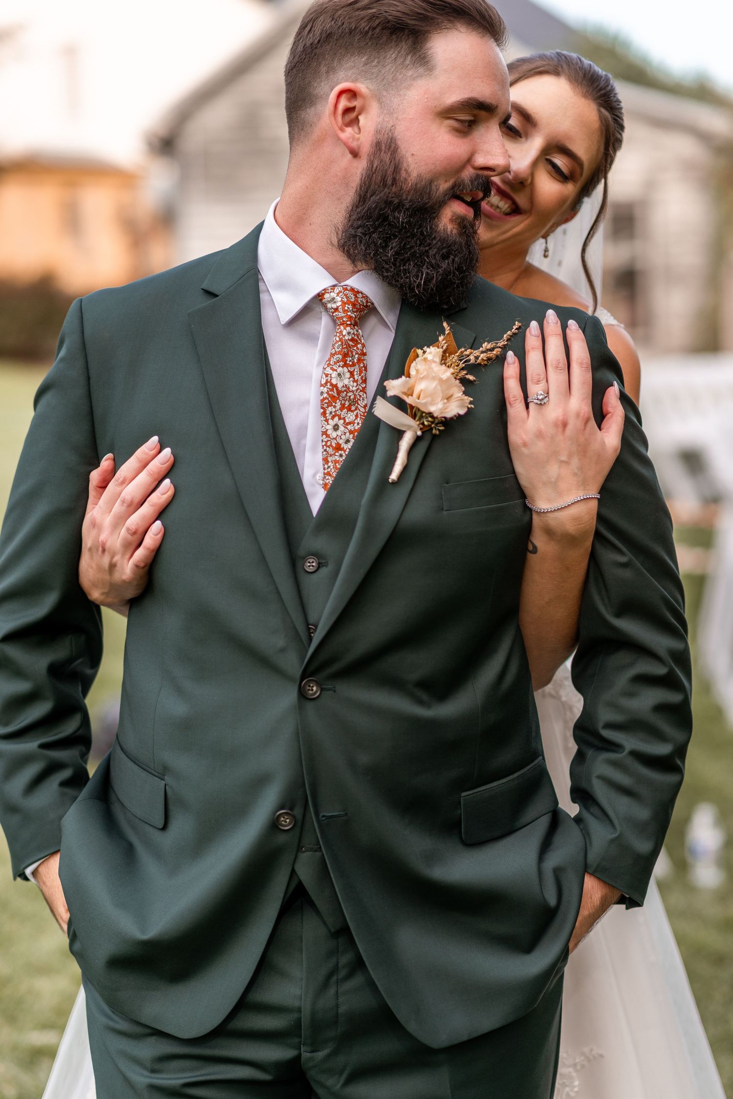 Bride embracing bearded groom from behind in dark green three-piece suit with floral boutonniere outdoors.
