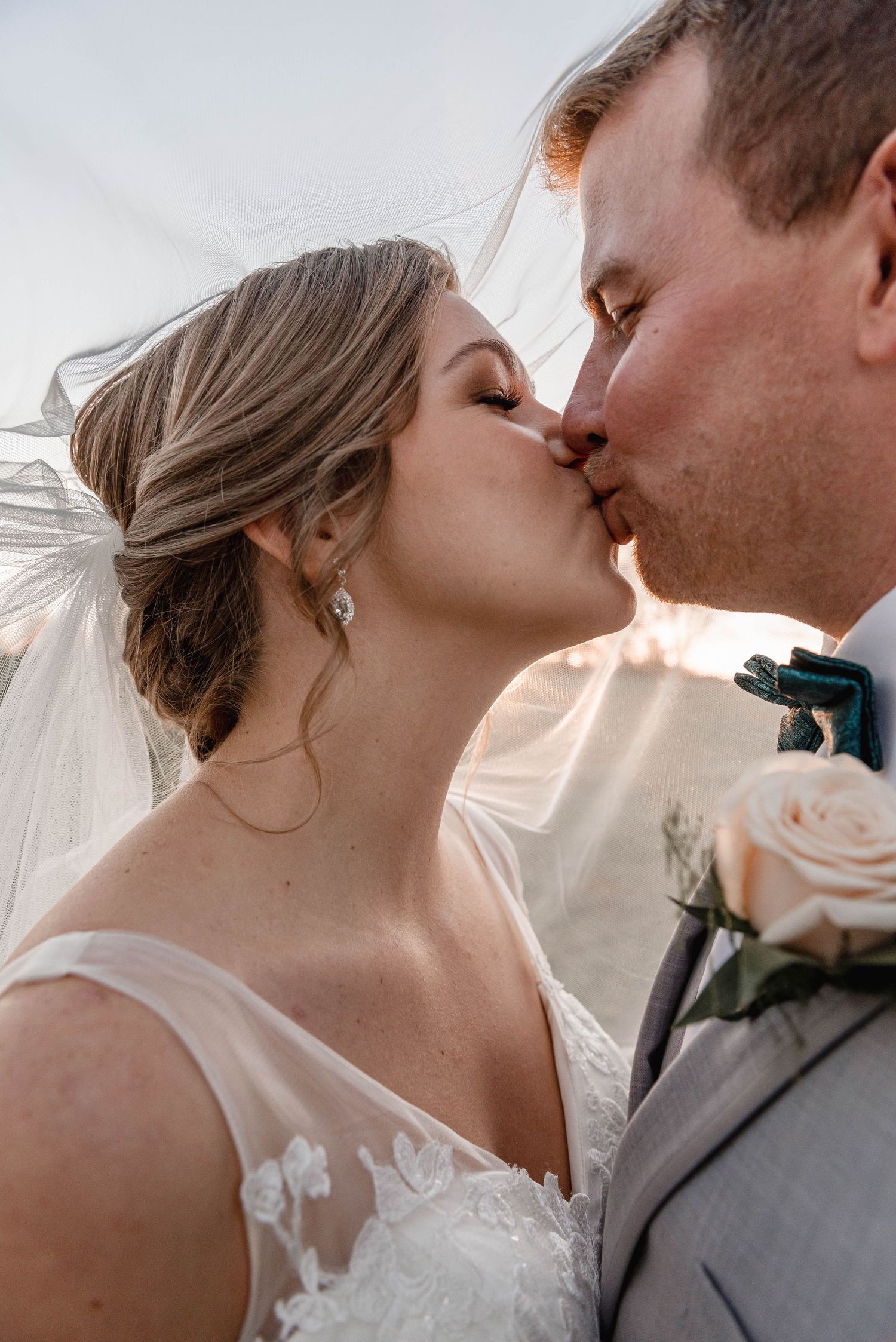Bride and groom sharing romantic kiss under flowing veil in golden outdoor light.
