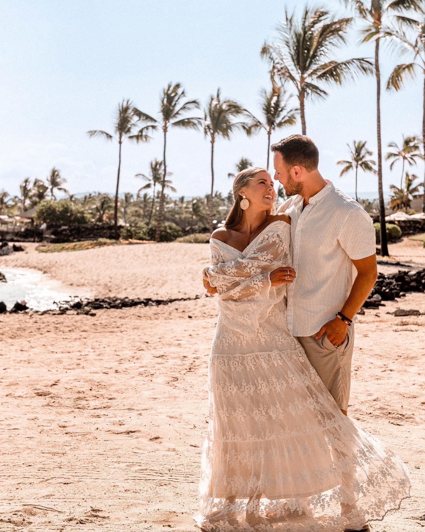 Couple in elegant attire sharing romantic moment on tropical beach with palm trees behind.
