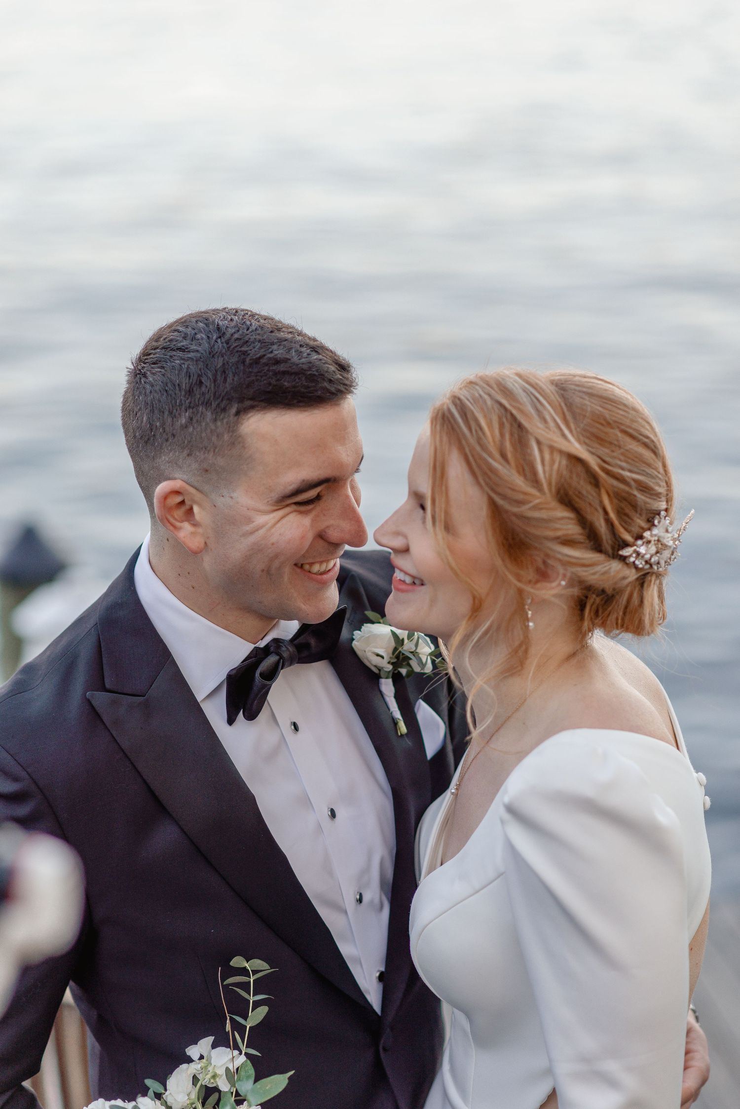 Smiling bride and groom in tuxedo share a romantic moment near the water at their wedding.