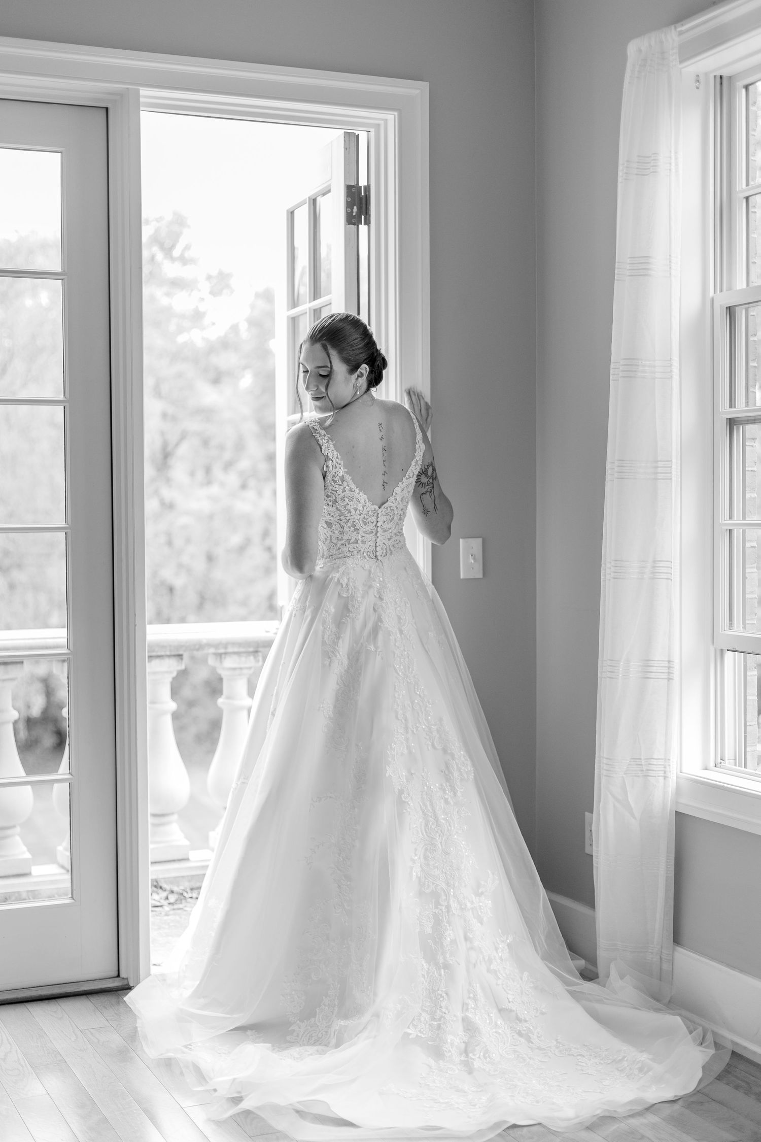 Elegant black and white portrait of a bride in a lace ball gown standing by French doors with flowing train.