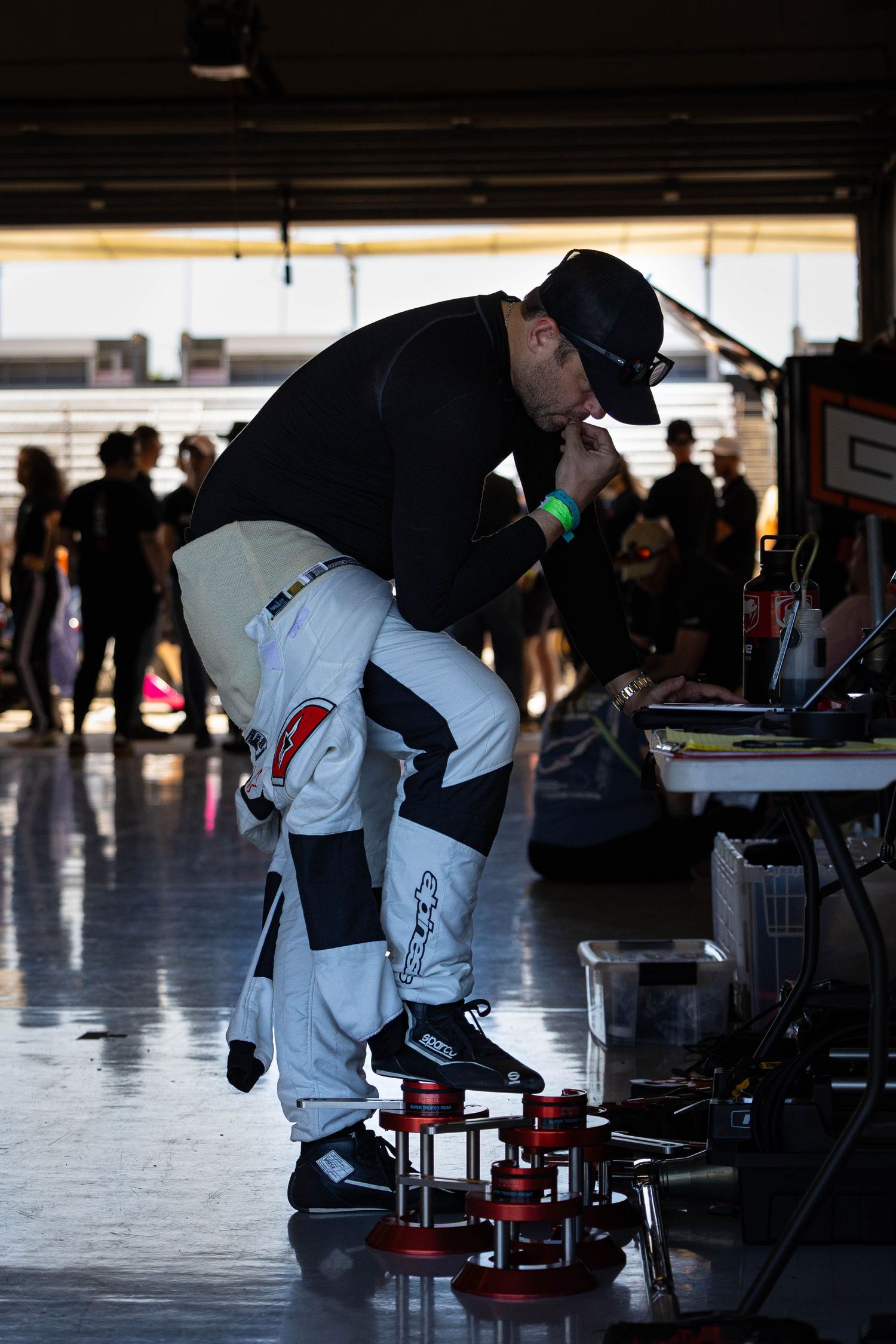 A mechanic in racing gear inspects equipment on the pit lane floor during a motorsport event.