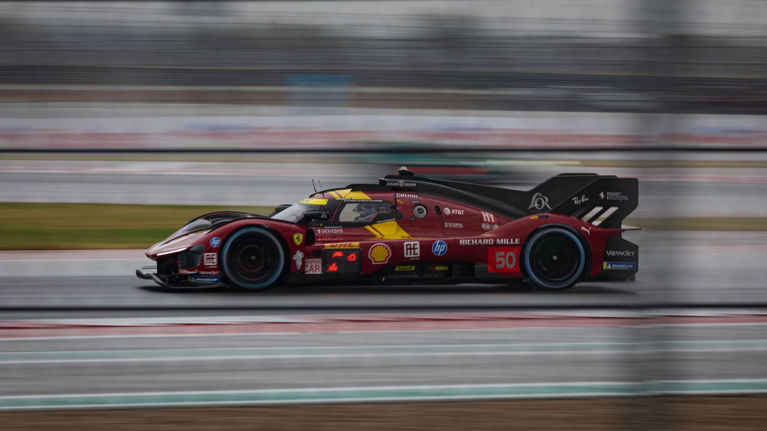 Ferrari LMH hypercar races on wet track with motion blur effect during endurance racing competition.