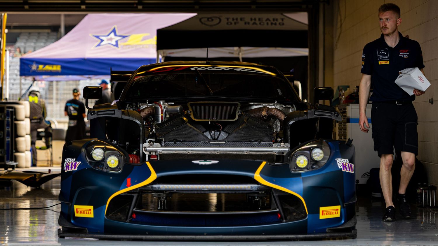 Aston Martin GT racing car parked in pit garage with team mechanics preparing for race day.