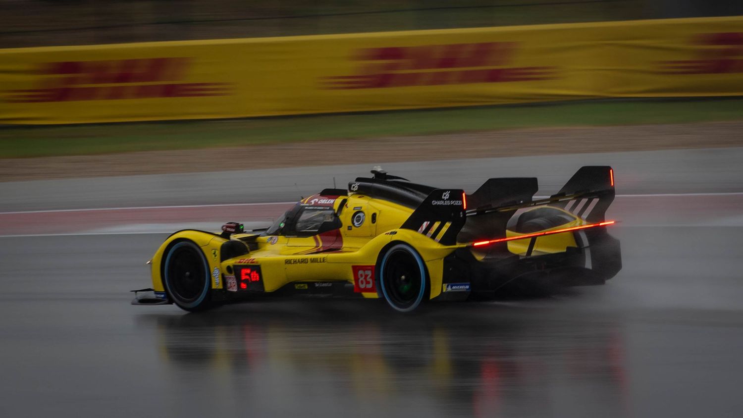 Yellow and black LMP prototype racing car speeding through wet track conditions with DHL advertising boards visible.