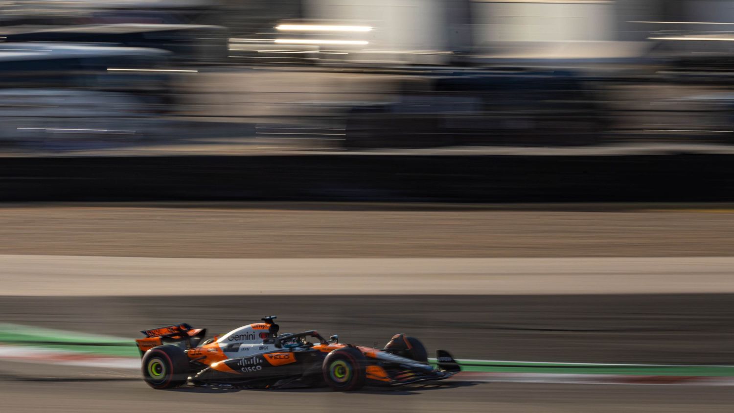 McLaren F1 car in papaya orange livery speeding along a racing circuit with grandstand motion blur.