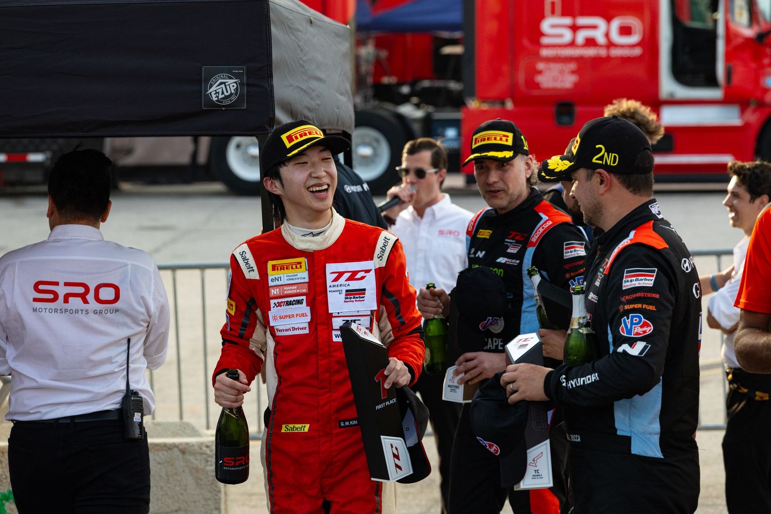 Racing drivers celebrate with champagne bottles at an SRO motorsport event podium ceremony.