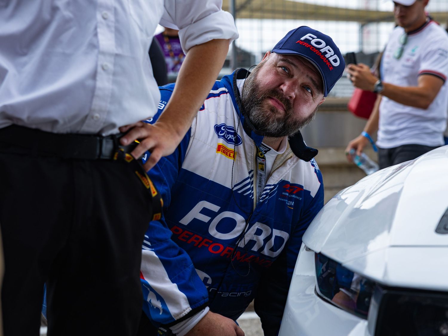 Ford Performance mechanic in blue racing suit inspects white race car during pit lane motorsport event.