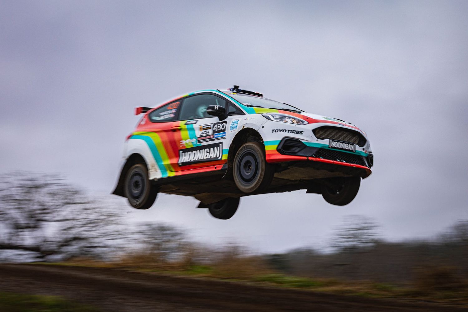 Rainbow-liveried Ford Fiesta rally car soaring through the air during a competitive rally stage.