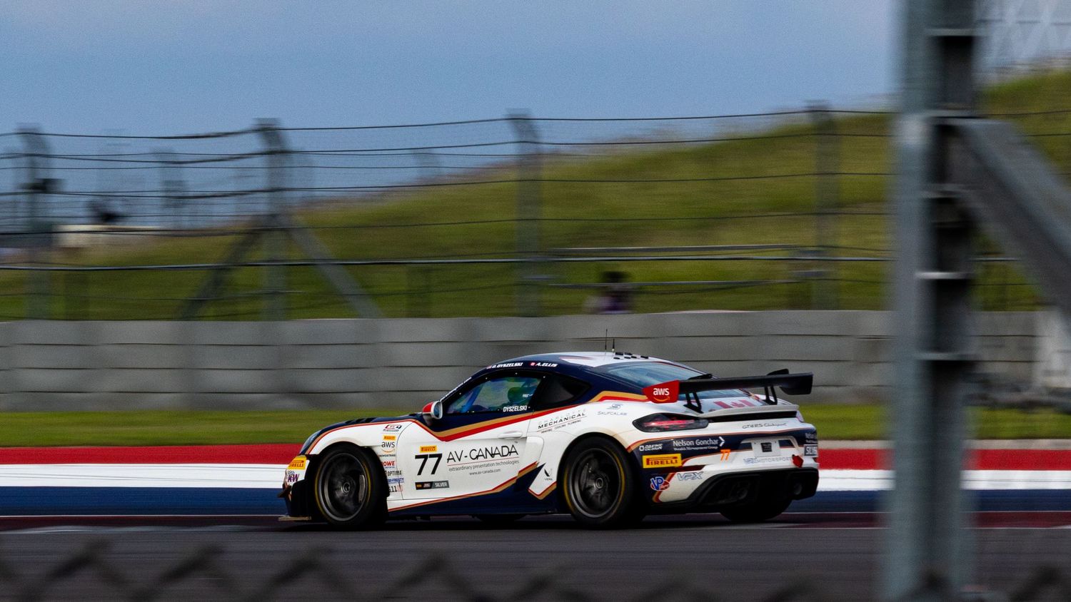 White number 77 GT race car navigates a turn at a motorsport circuit during a GT racing event.