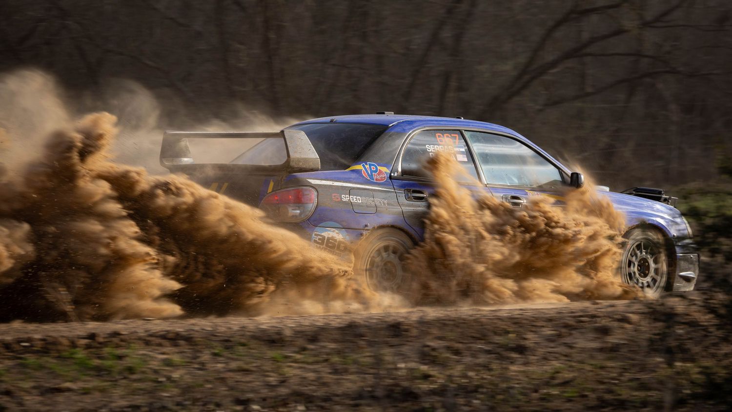 Blue rally car sliding aggressively through muddy terrain sending dirt flying on a forest stage.
