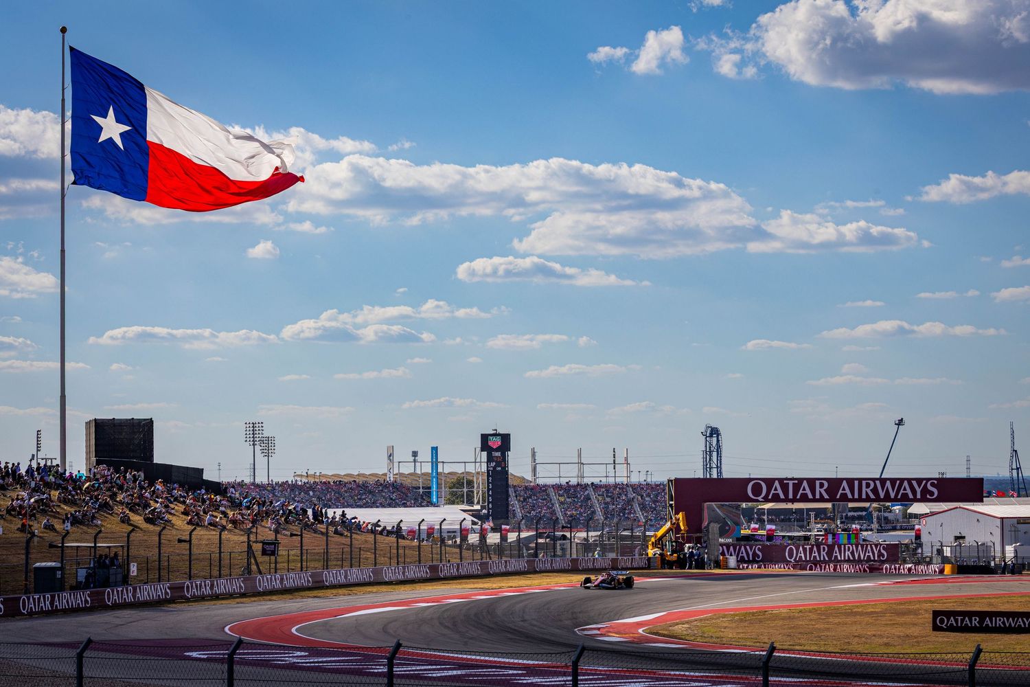 Texas flag waves over Circuit of the Americas Formula 1 race track with Qatar Airways signage under blue skies.