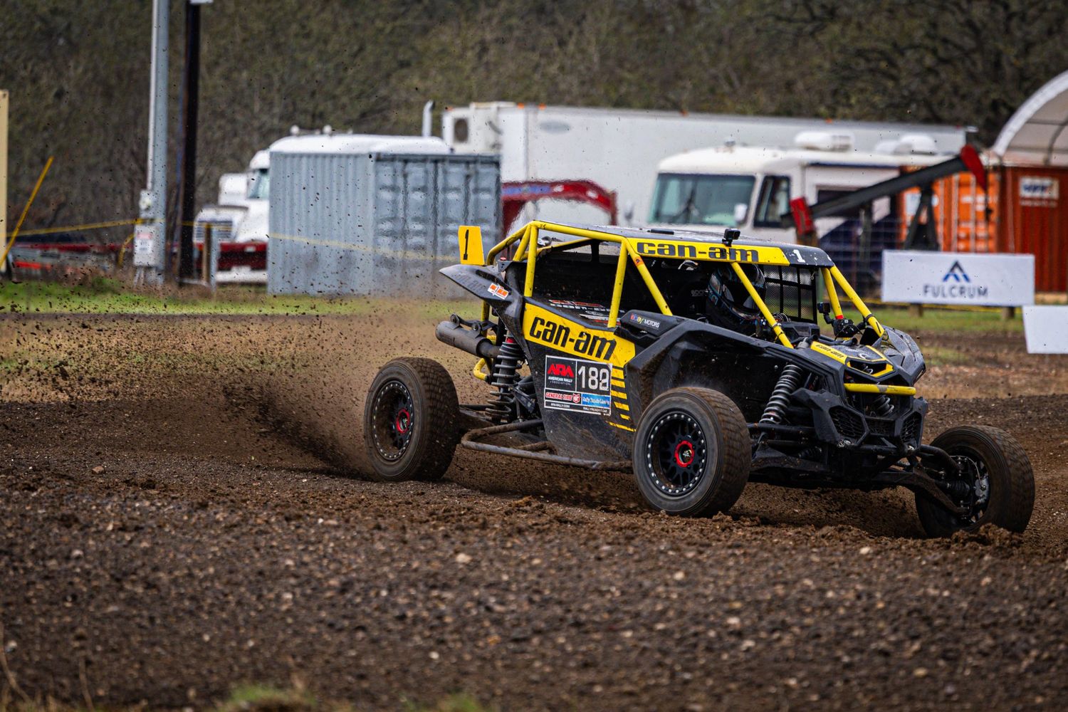 Can-Am side-by-side UTV racing through muddy dirt course at off-road rally event.