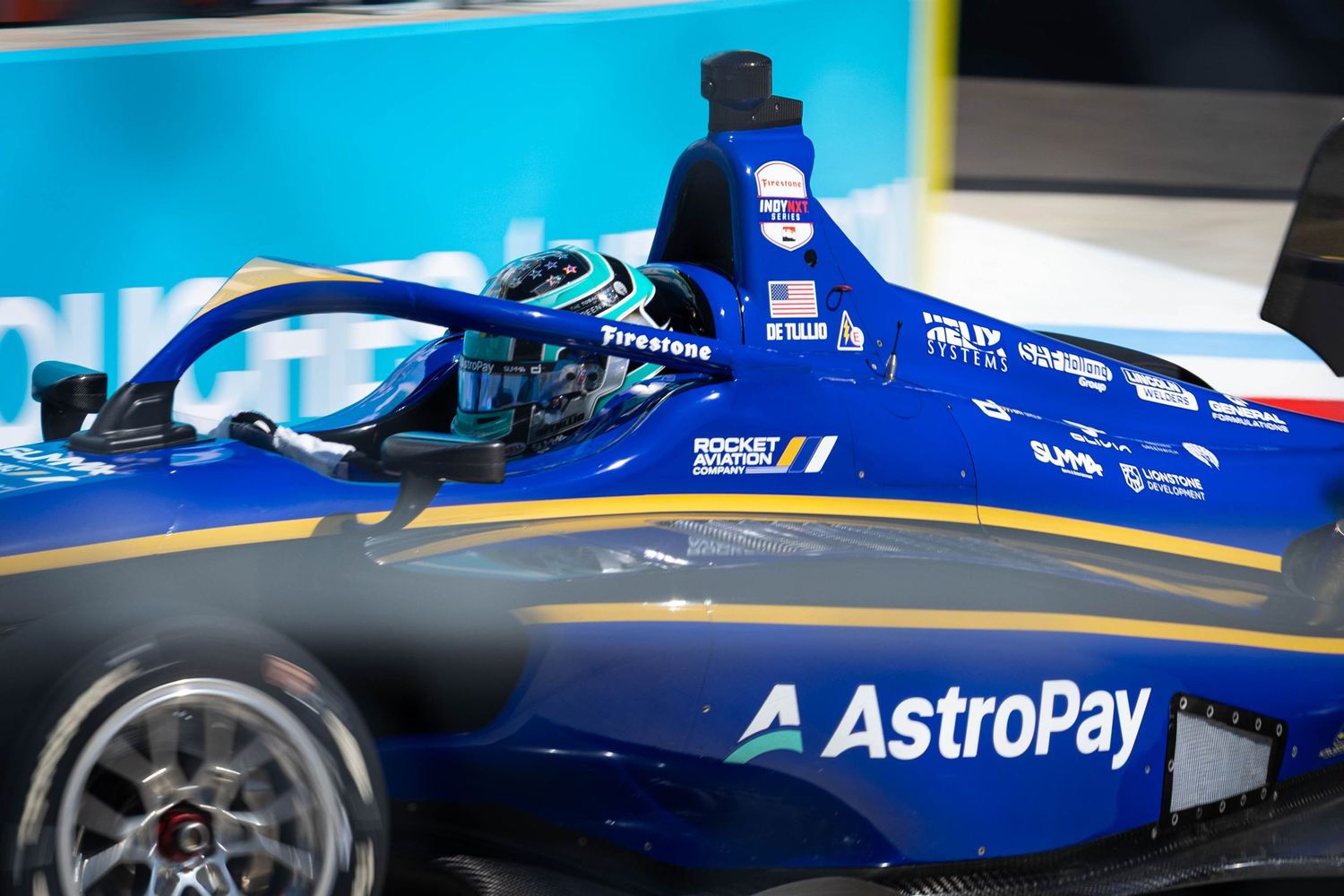 Blue AstroPay sponsored IndyCar single-seater racing at speed showing cockpit detail with Firestone tire branding.