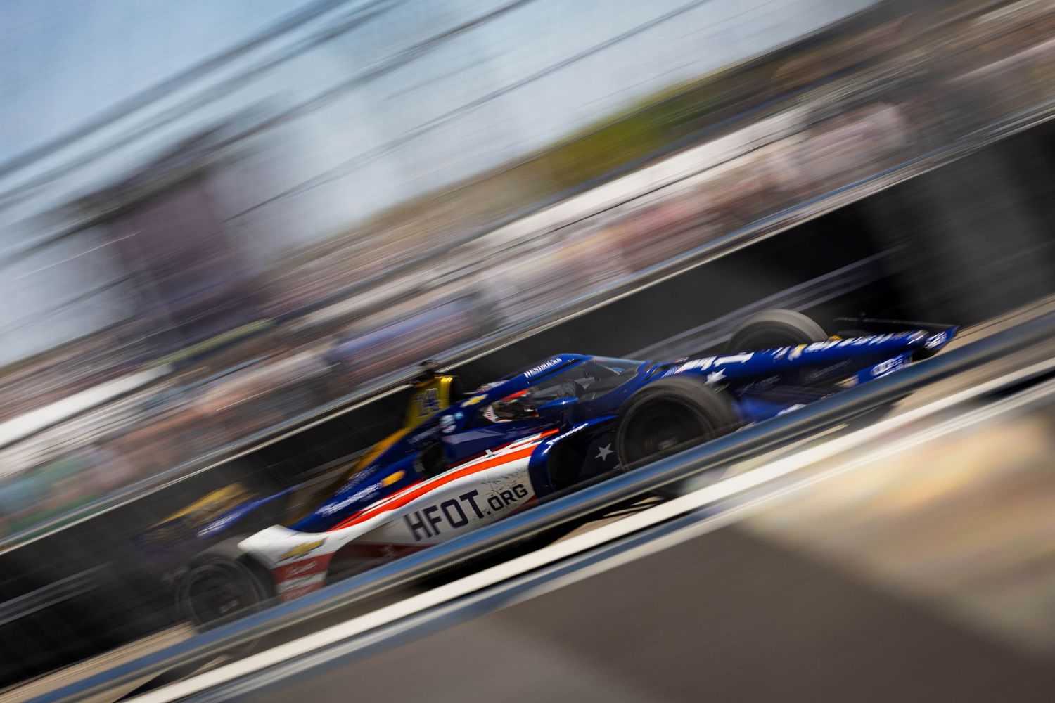 Blue IndyCar with HFOT sponsor livery racing at high speed through a motorsport circuit pit straight.