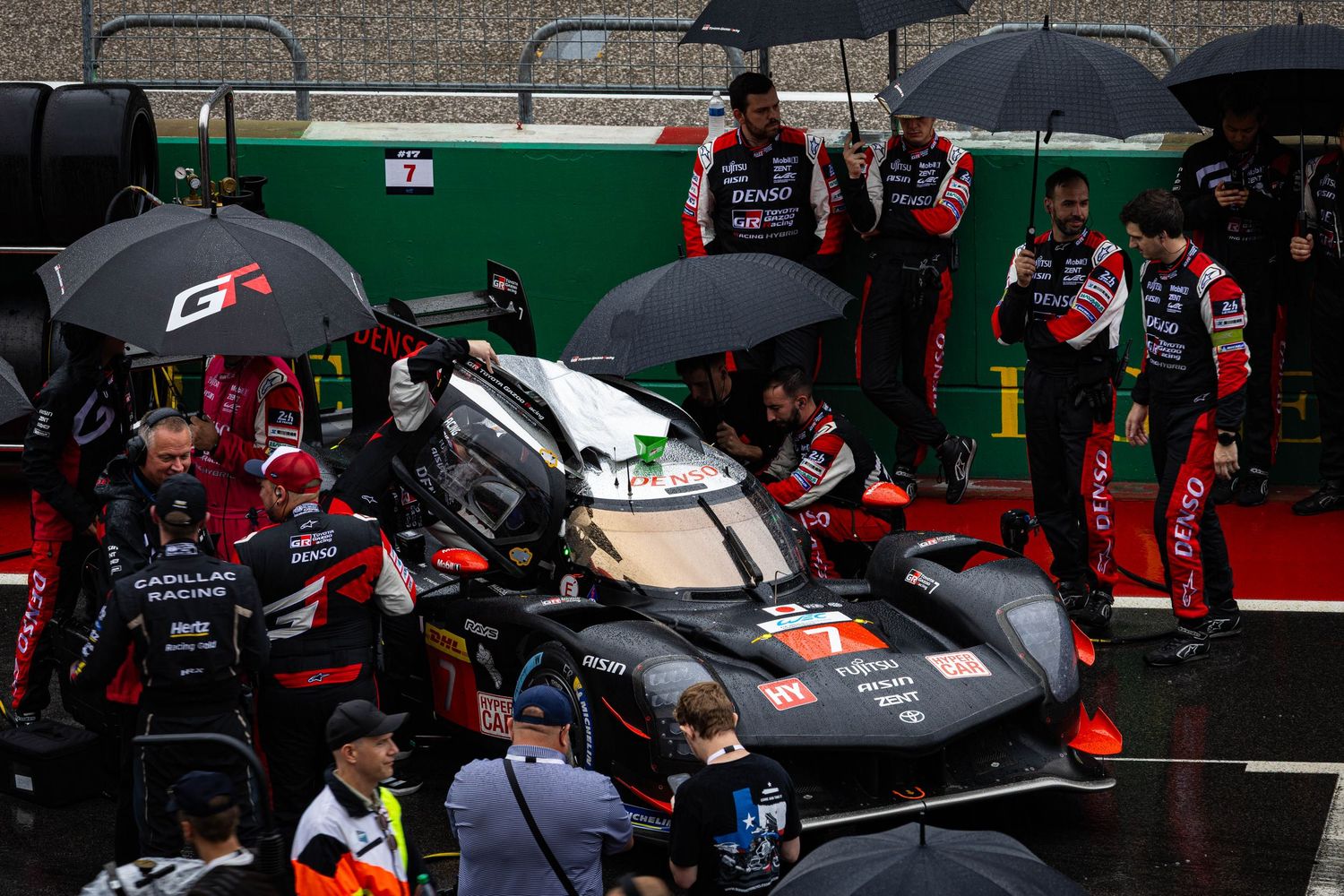 Toyota GR racing team mechanics working on a prototype endurance race car in wet pit lane conditions under umbrellas.