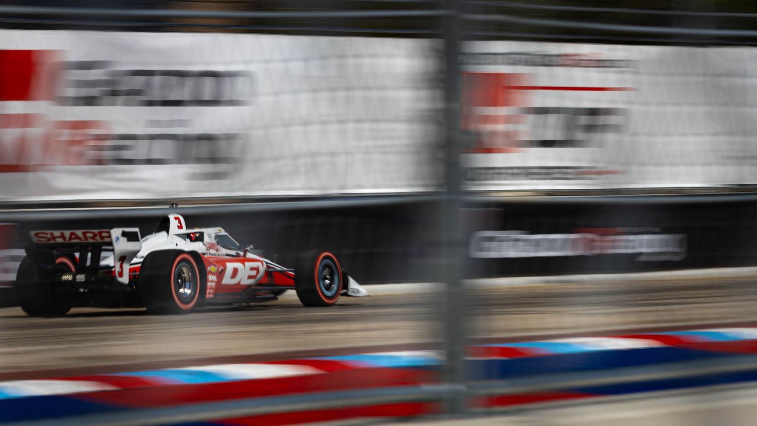 IndyCar racing past sponsor banners on street circuit with motion blur effect.