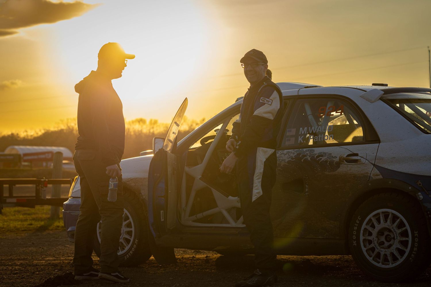 Two silhouetted figures conversing beside a rally car during a dramatic golden sunset at a racing event.