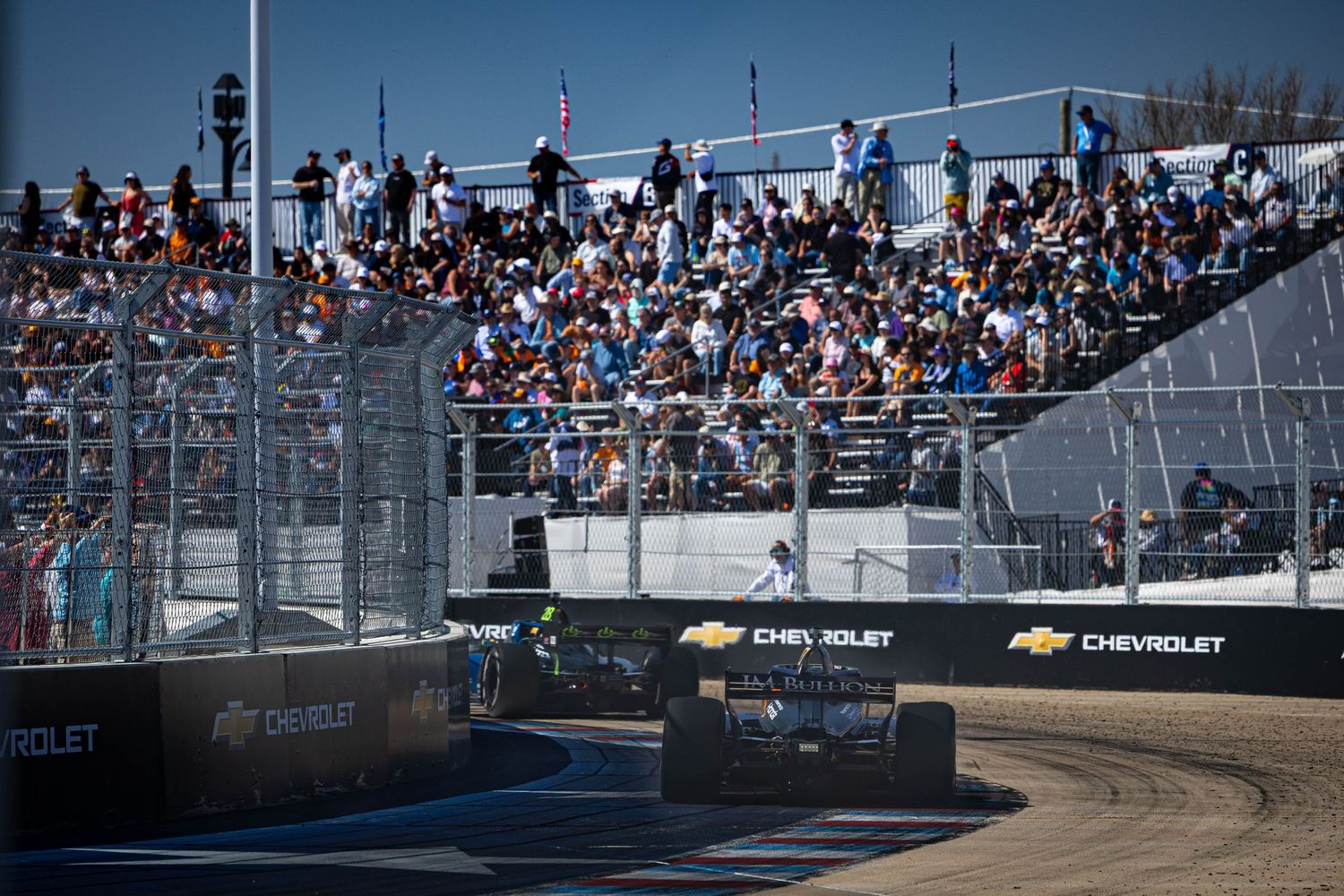 IndyCar race car speeds past packed grandstands at a street circuit with Chevrolet sponsorship banners on a sunny day.