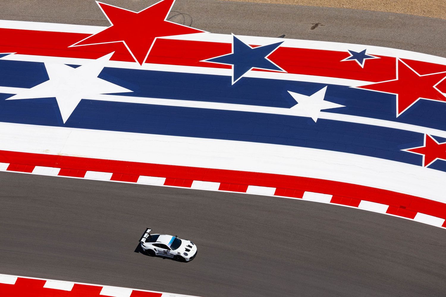 White GT race car navigates circuit beneath giant patriotic American stars and stripes track mural.