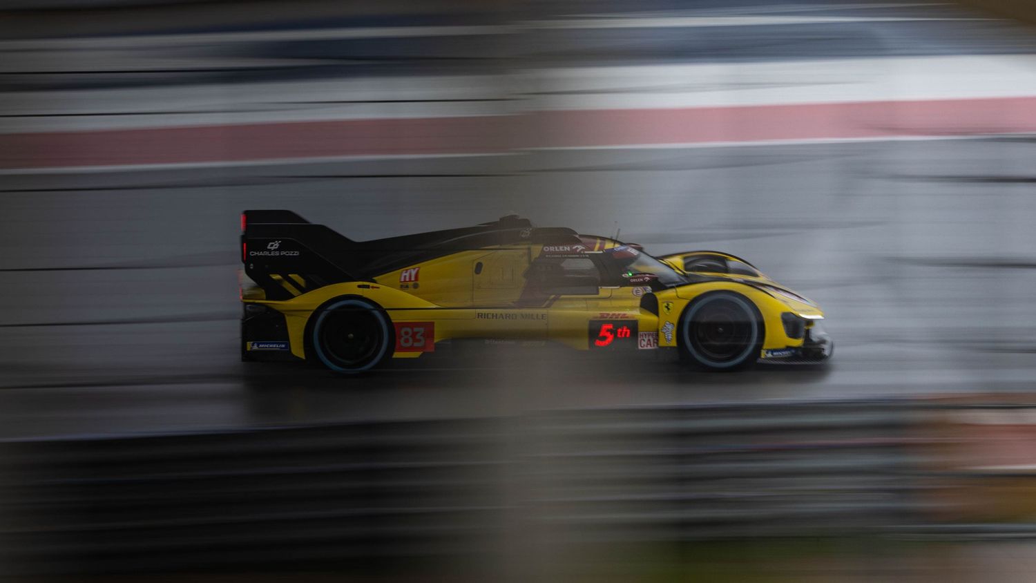 Yellow Corvette GTP prototype race car speeds along wet racing circuit with motion blur effect.