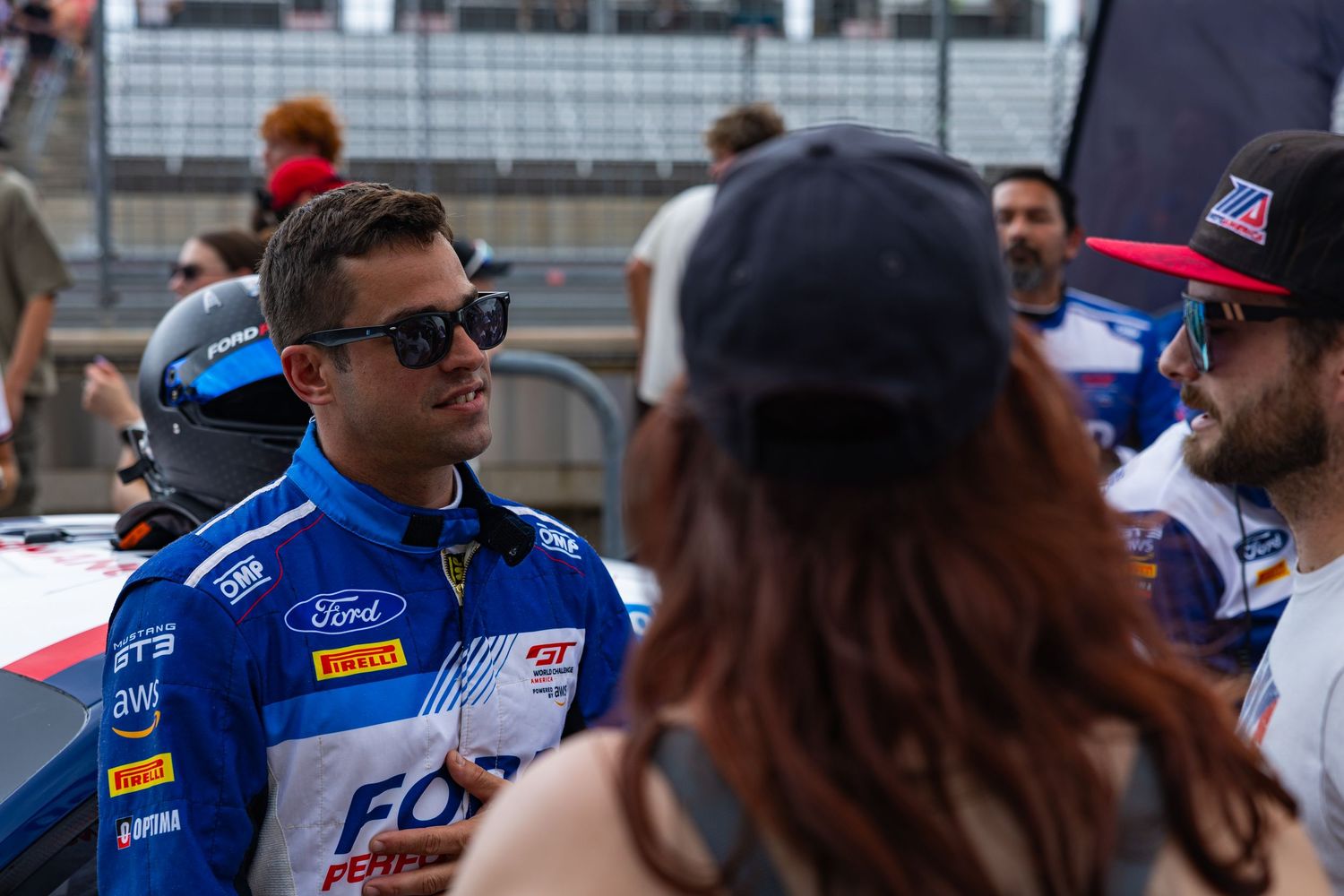 A Ford Performance GT3 racing driver in blue uniform converses with crew members on the grid.