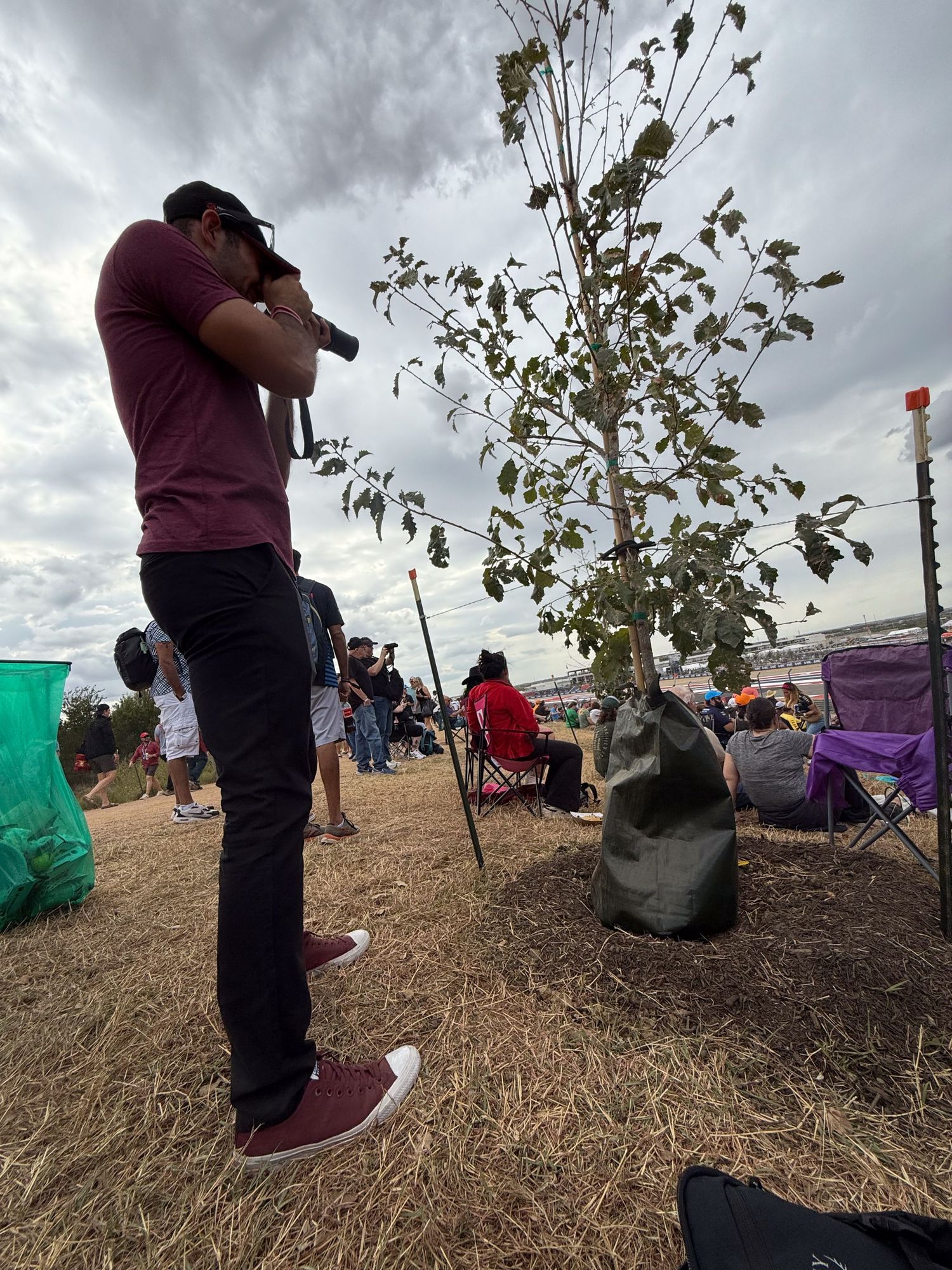 Photographer shooting with camera at outdoor motorsport event beside small tree.