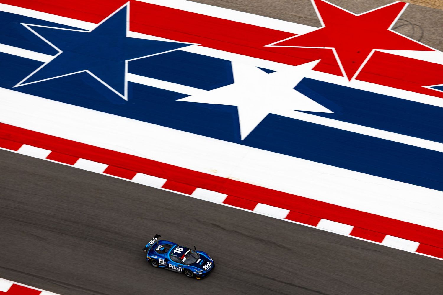 A blue GT racing car speeds past a patriotic American stars and stripes mural on a race circuit.